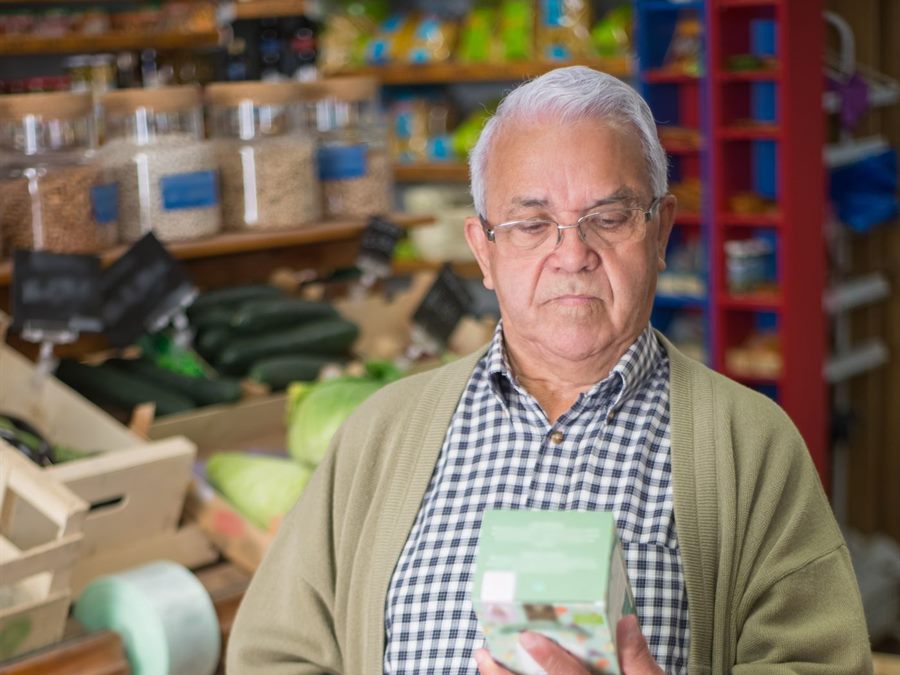 Homem de óculos parado no corredor do mercado lendo o rótulo de uma embalagem